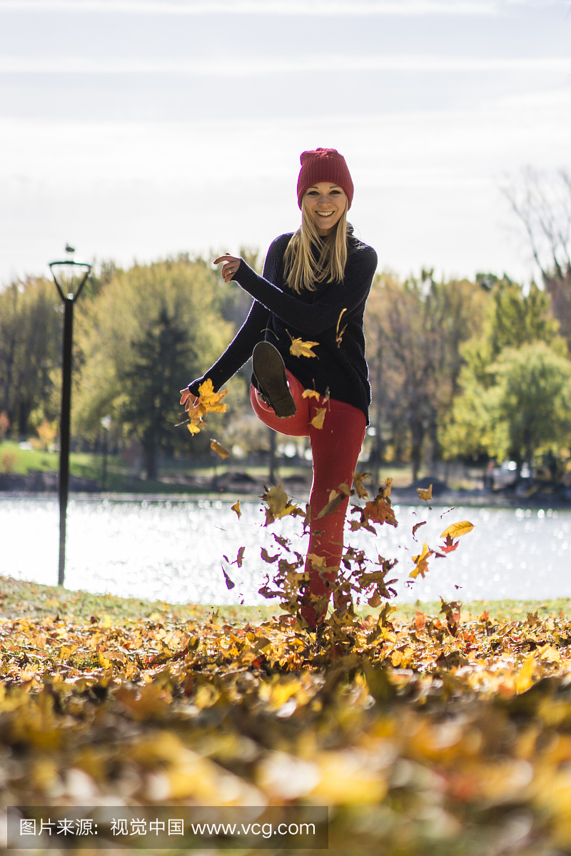 穿着红色编织帽子的愉快的少妇在公园里踢秋天