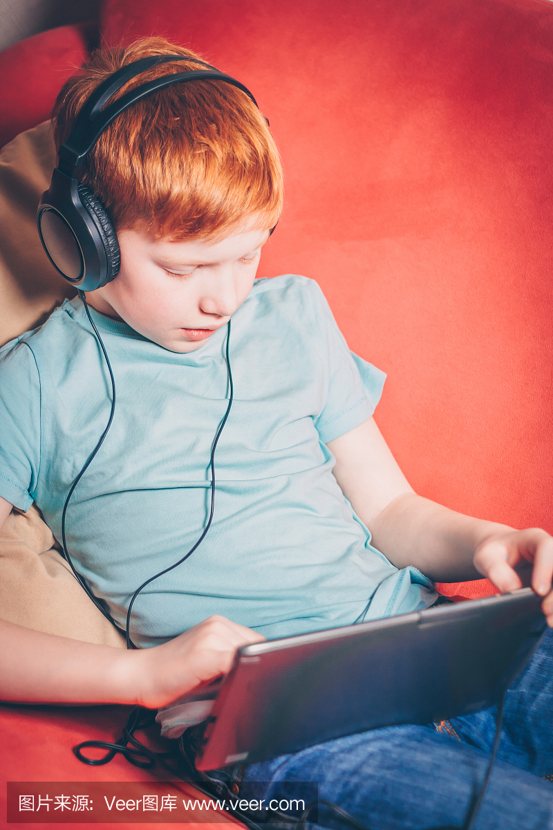 红头发的男孩坐在沙发上在平板电脑上玩游戏