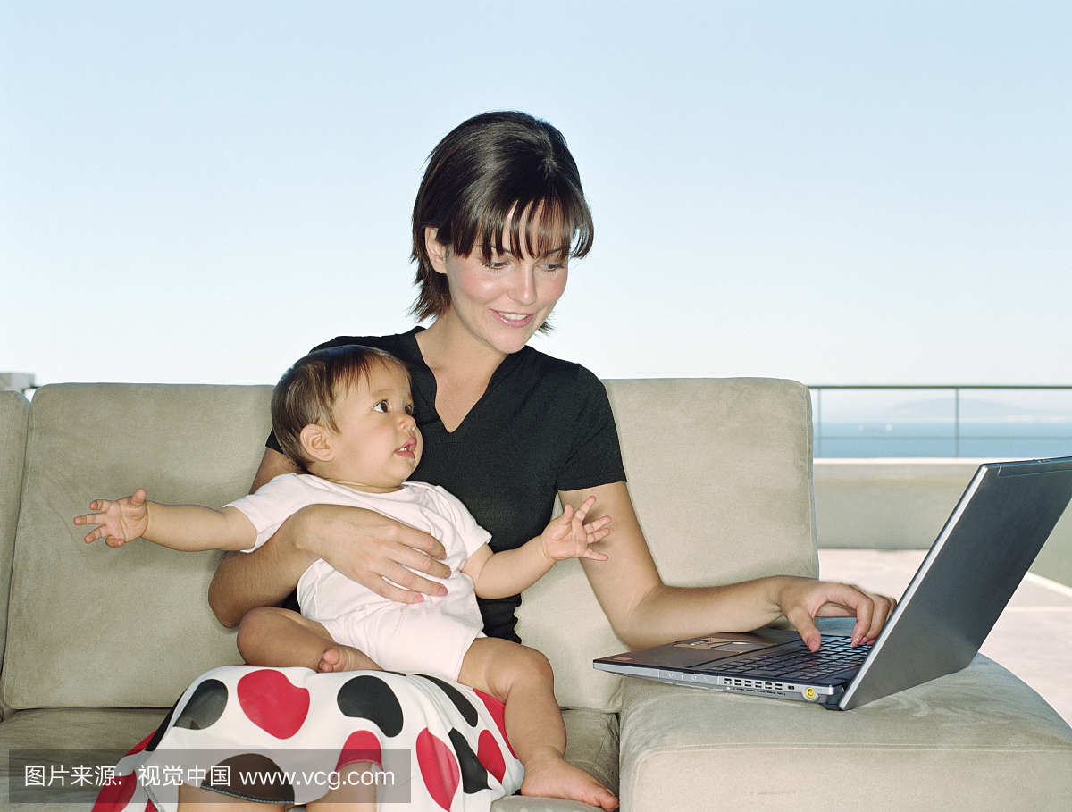 母亲抱着女婴(6-9个月)在沙发上,妈妈用笔记本