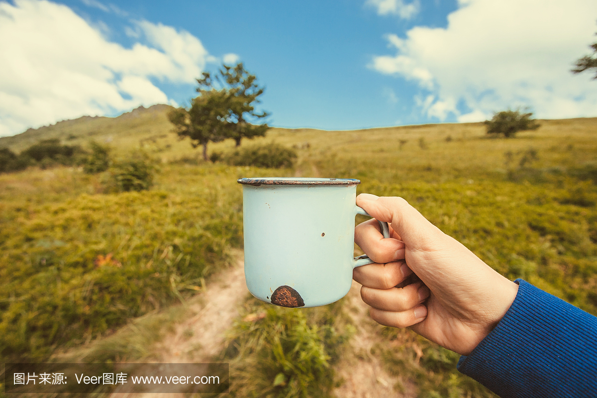 金属杯与茶在手上的旅游者在白色云彩下的山上