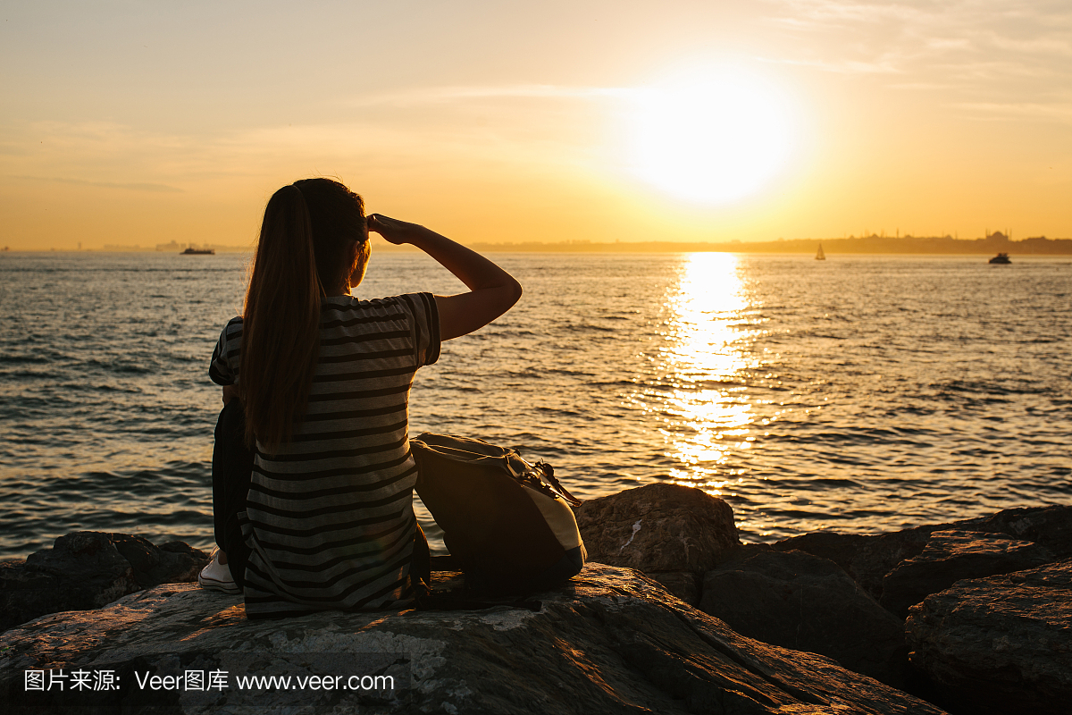 一个年轻的旅游女孩背着背包坐在海边的岩石上